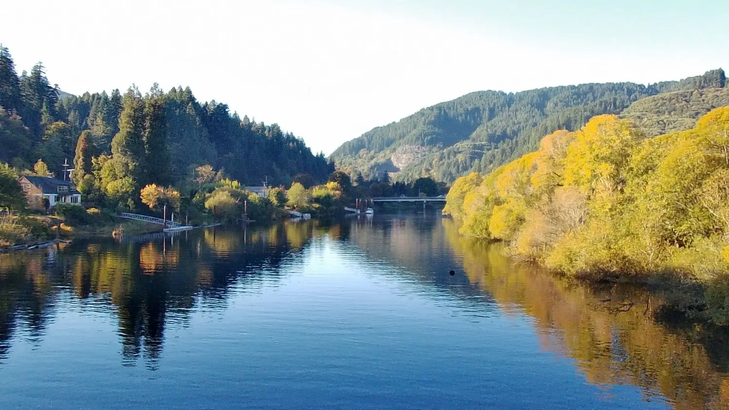 A wide river reflects a clear sky, flanked by lush green and golden autumn trees, with distant hills and a bridge.