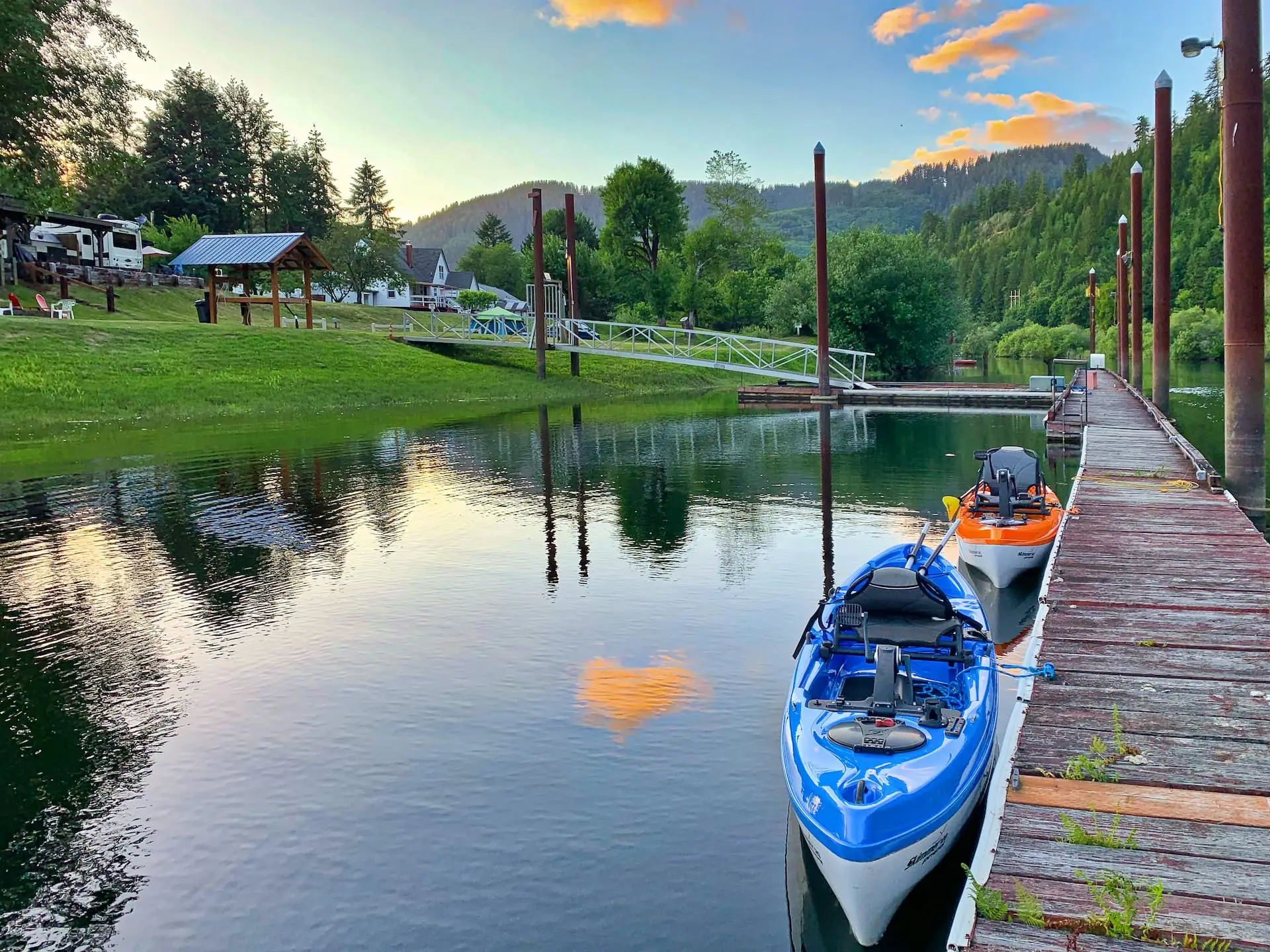 Two kayaks, one blue and one orange, are docked at a wooden marina on a calm river with green hills and trees under a sunset sky.