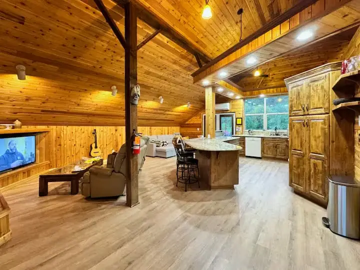 A spacious, rustic-style living area with wood-paneled walls and ceiling, a kitchen island, and a TV showing a man.