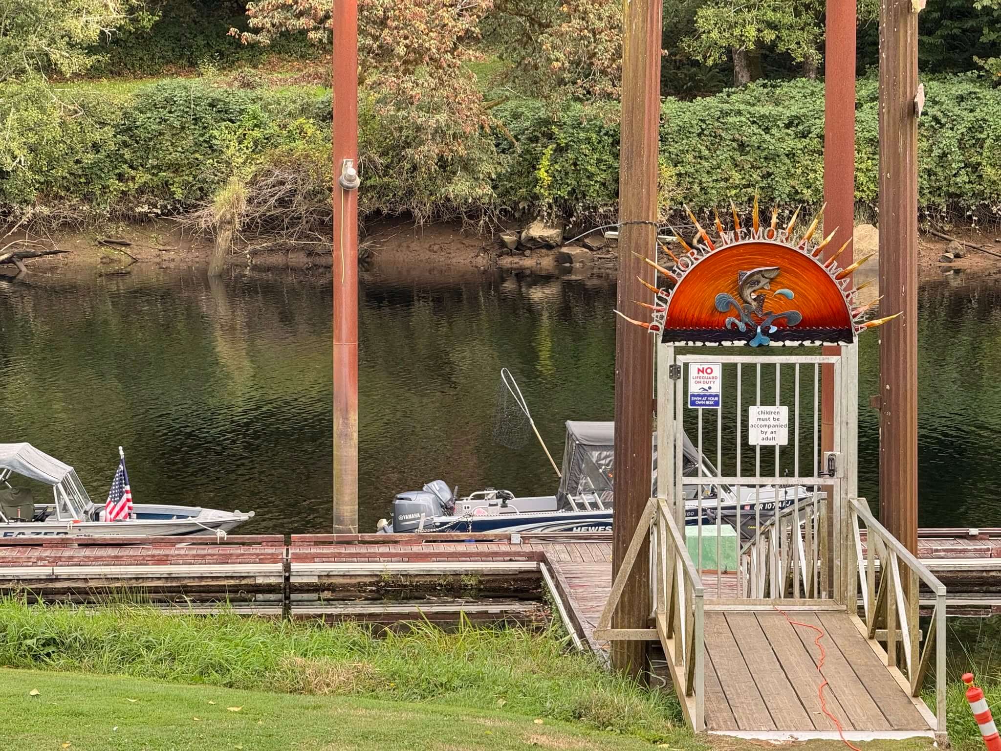 A marina with two boats docked on a calm river, surrounded by lush green trees. A metal gate with a sun and fish design stands at the entrance.