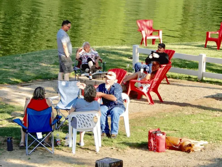 A group of people relaxing in chairs by a calm green body of water, with a dog sleeping nearby on the grass.