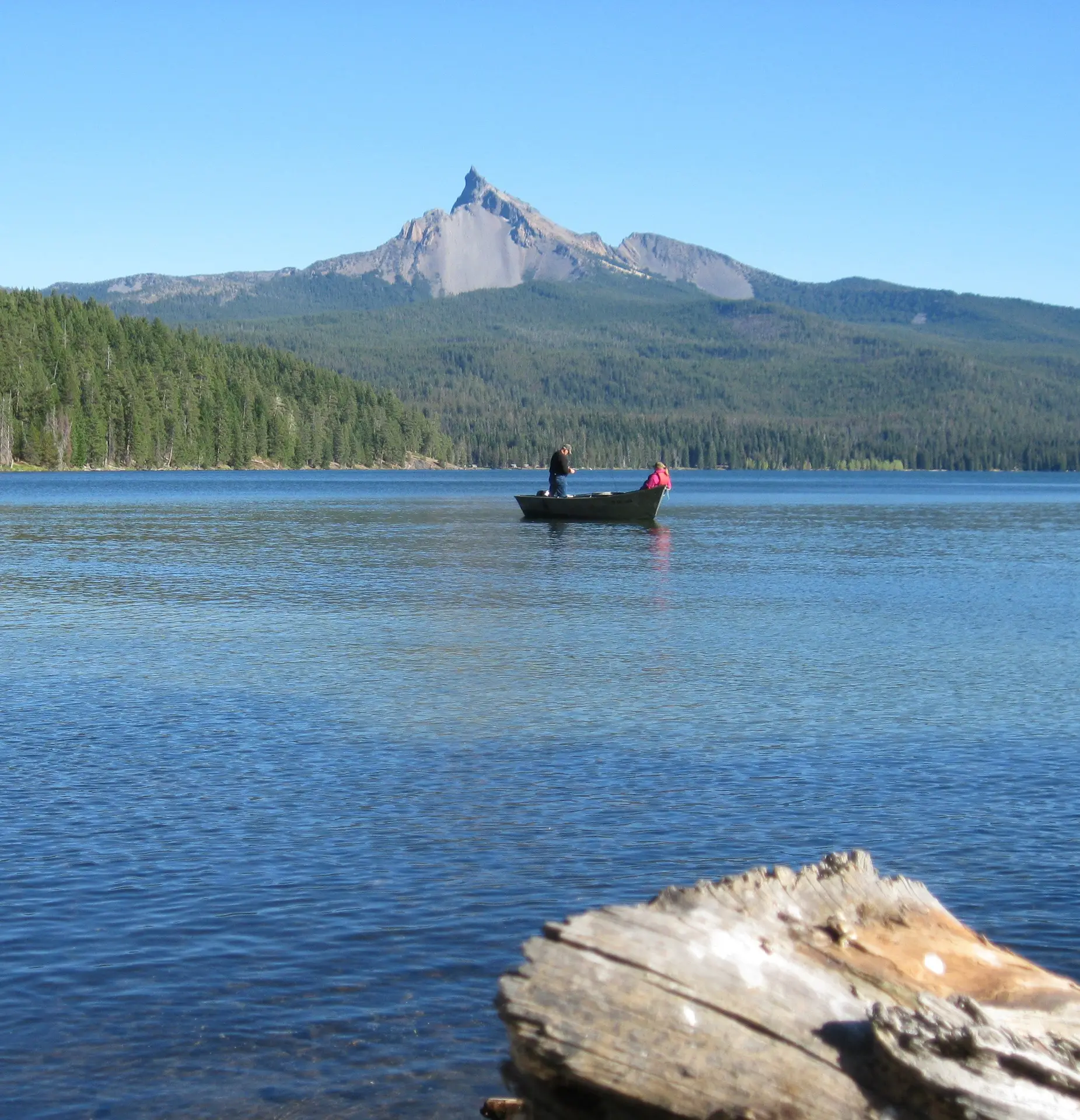 Two people fish from a small boat on a calm lake with a large, pointed mountain and a dense forest in the background.