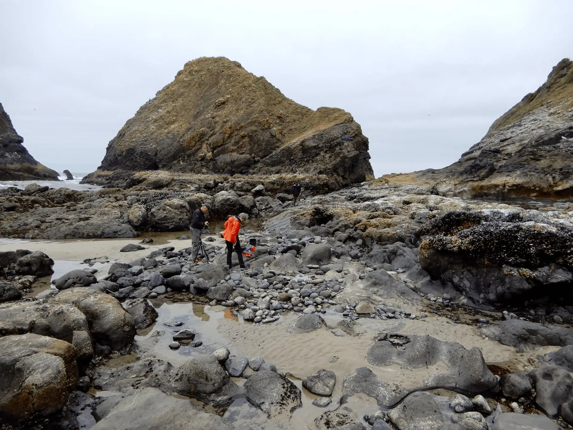 Two people explore a rocky beach with large boulders and sandy patches under an overcast sky, with a massive hill in the background.