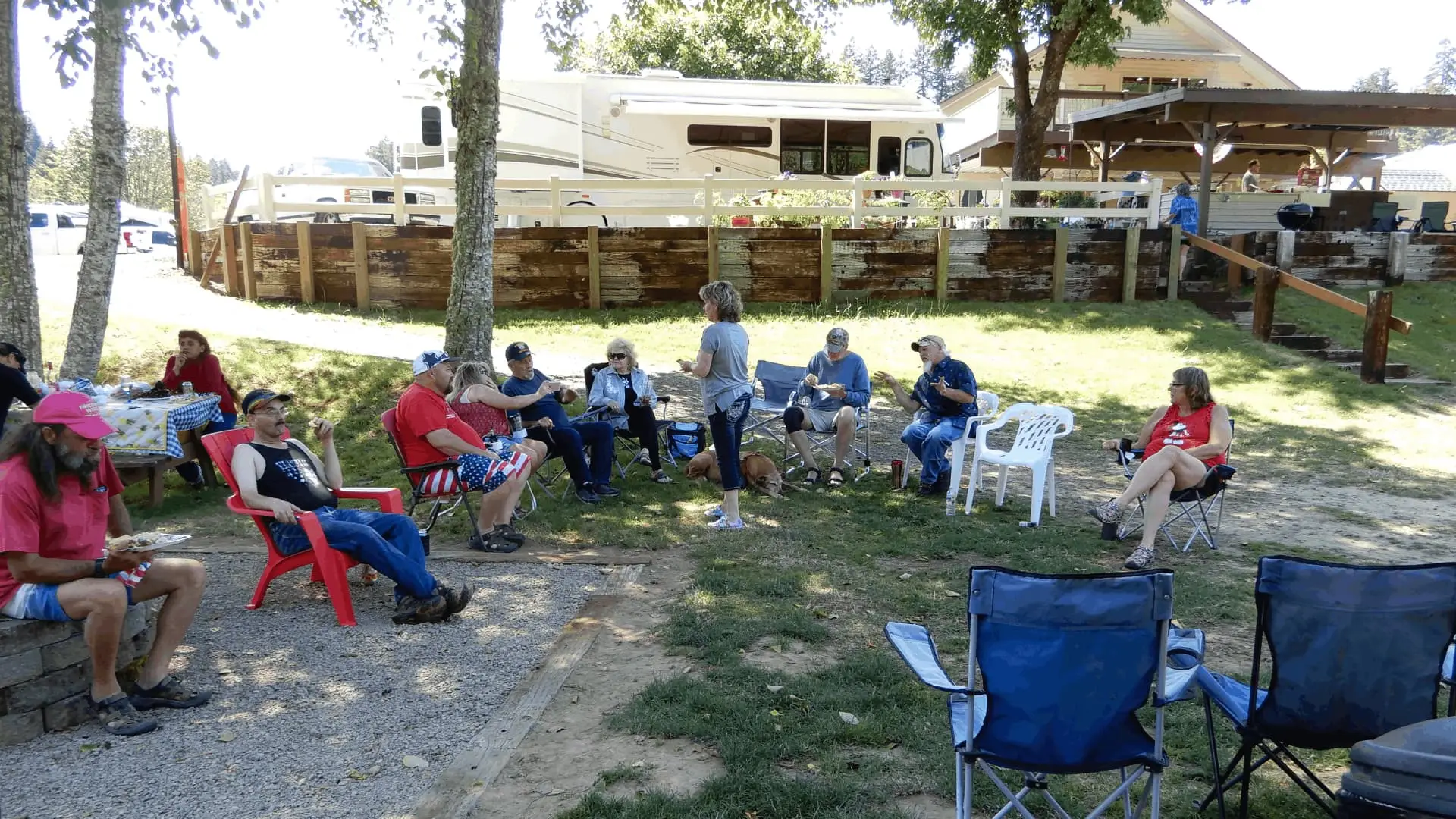People gather in a circle outdoors, some seated in chairs, others standing, with an RV and building in the background.