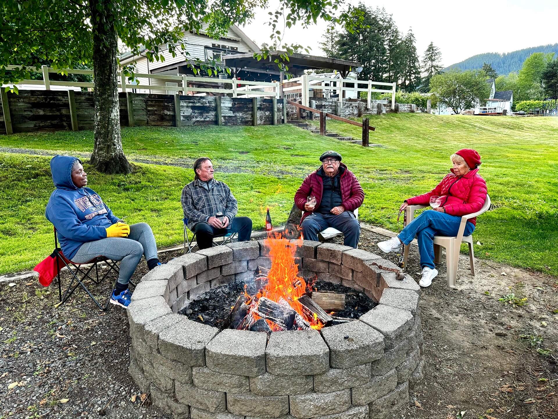 Four adults sit around a stone fire pit with a roaring fire, enjoying drinks and conversation on a grassy lawn.