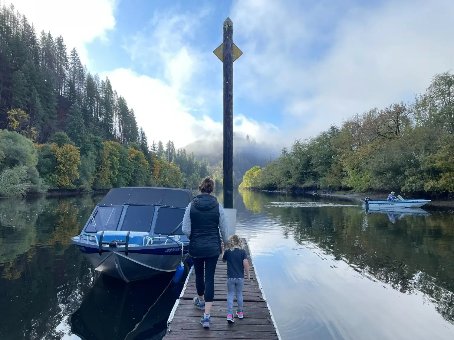 A woman and a child walk down a wooden dock towards a river with boats, surrounded by lush green and autumn trees.