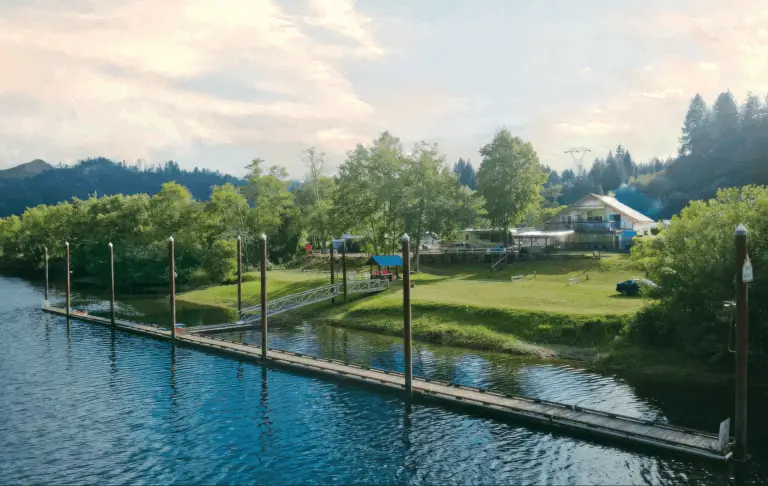 An aerial view of Maple Lane RV Park and Marina on the Oregon Coast, featuring a long wooden dock extending into calm blue water. Lush green trees and a building are visible on the grassy shore under a partly cloudy sky.