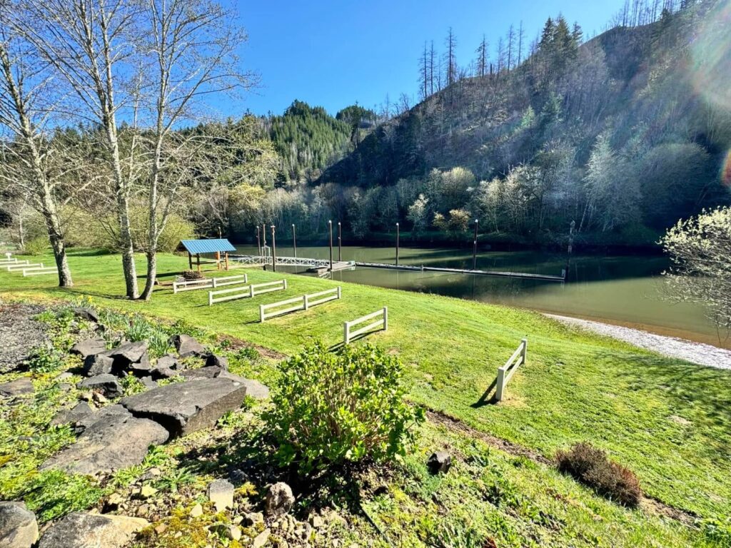 A sunny view of Maple Lane RV Park and Marina, east of Florence on the Oregon Coast, showing a green lawn, trees, and a dock extending into calm water.