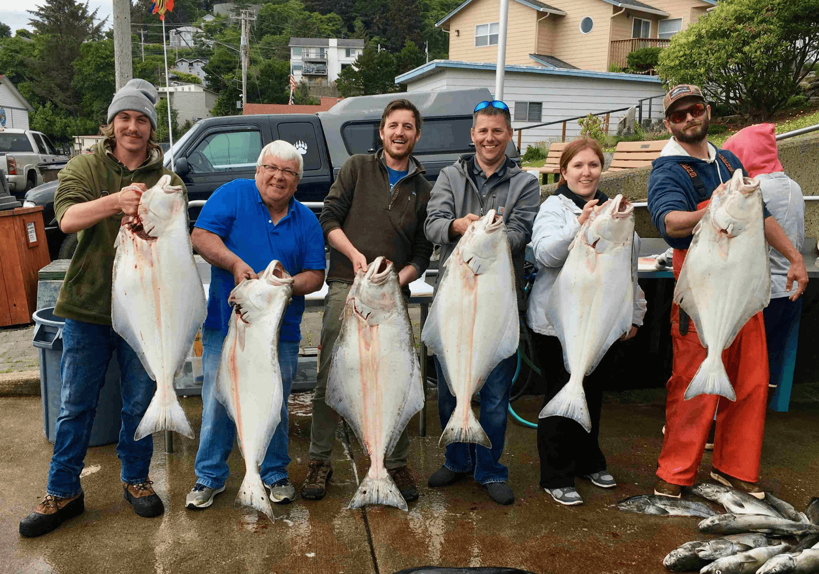 Six smiling people, four men and two women, proudly display their large halibut catches in a coastal town setting.