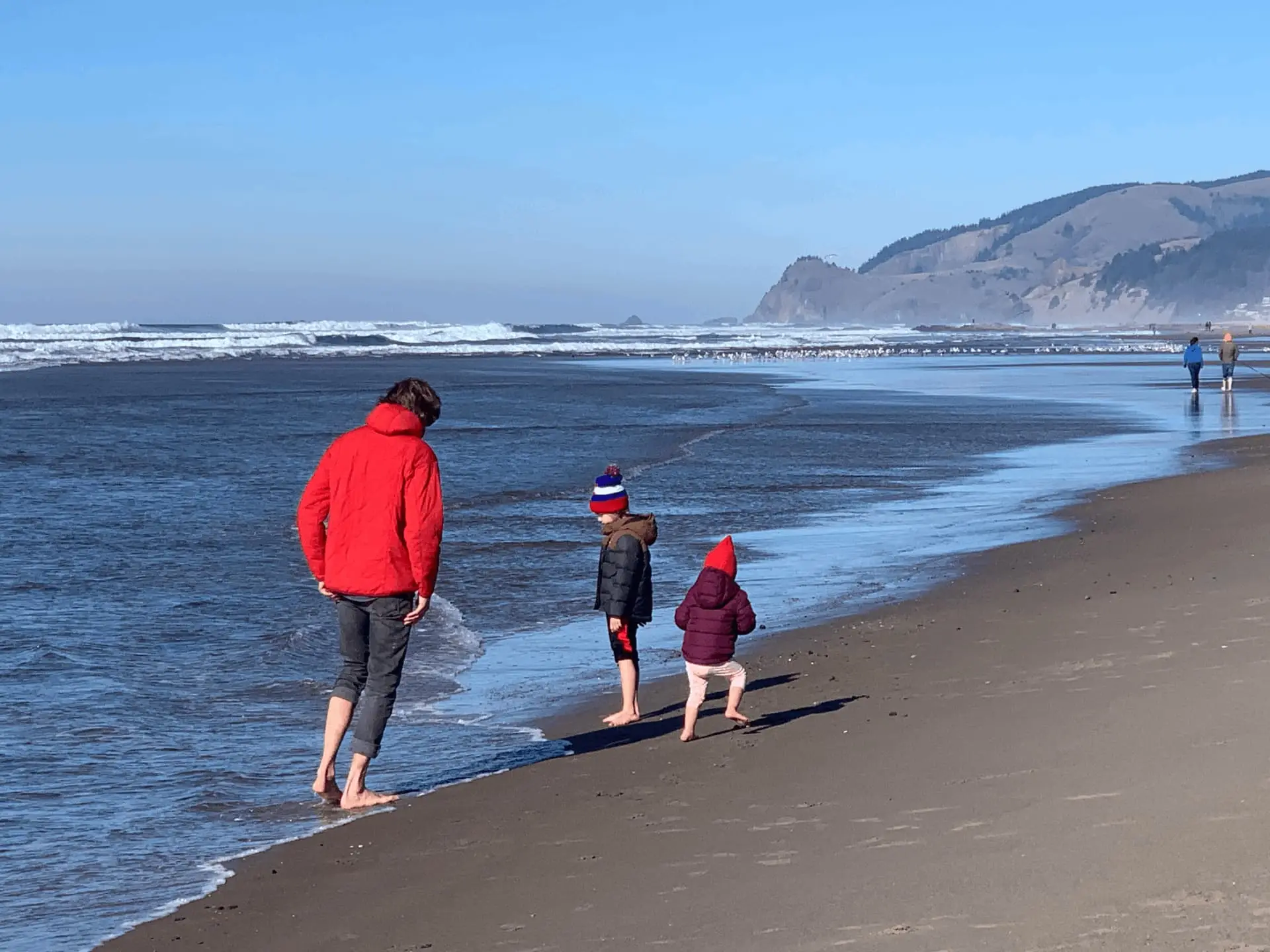 A man in a red jacket and two children in winter hats walk barefoot along the wet sand of a beach on the Oregon Coast.