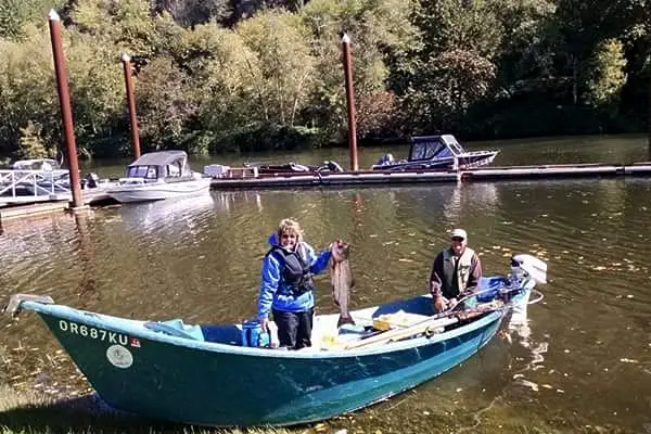 A woman in a life vest holds two fish while standing in a small boat next to a man, with a marina and trees in the background.