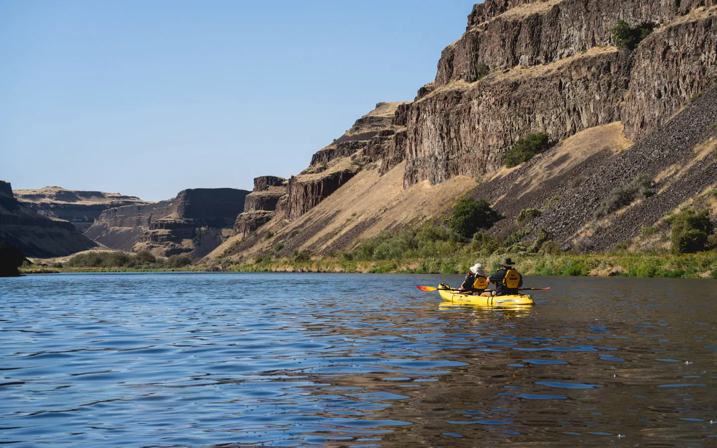 Three people in a yellow kayak paddle on a wide river with steep, rocky, and dry hillsides under a clear blue sky.