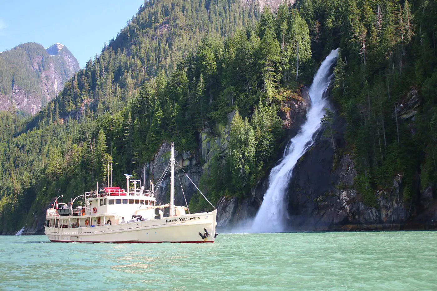 A large white boat, the Pacific Yellowfin, cruises on turquoise water next to a lush green mountain with a powerful waterfall.
